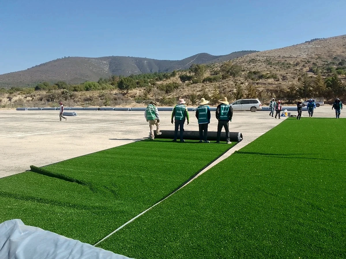 Equipo de Sportmaster extendiendo rollos de pasto sintético sobre la base del campo de béisbol en Santiago Tenango, Puebla