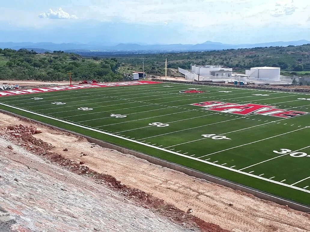 Campo de fútbol americano con pasto sintético en Arkansas State University Campus Querétaro, con la zona de anotación Arkansas State y los edificios del campus