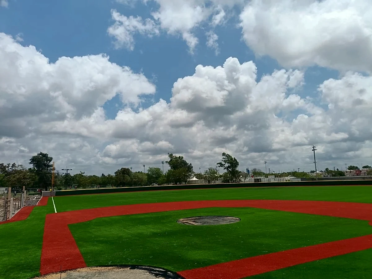 Campo de béisbol con pasto sintético verde y zonas en pasto rojo bajo un cielo con nubes en el Parque Felipe Braza, Matamoros